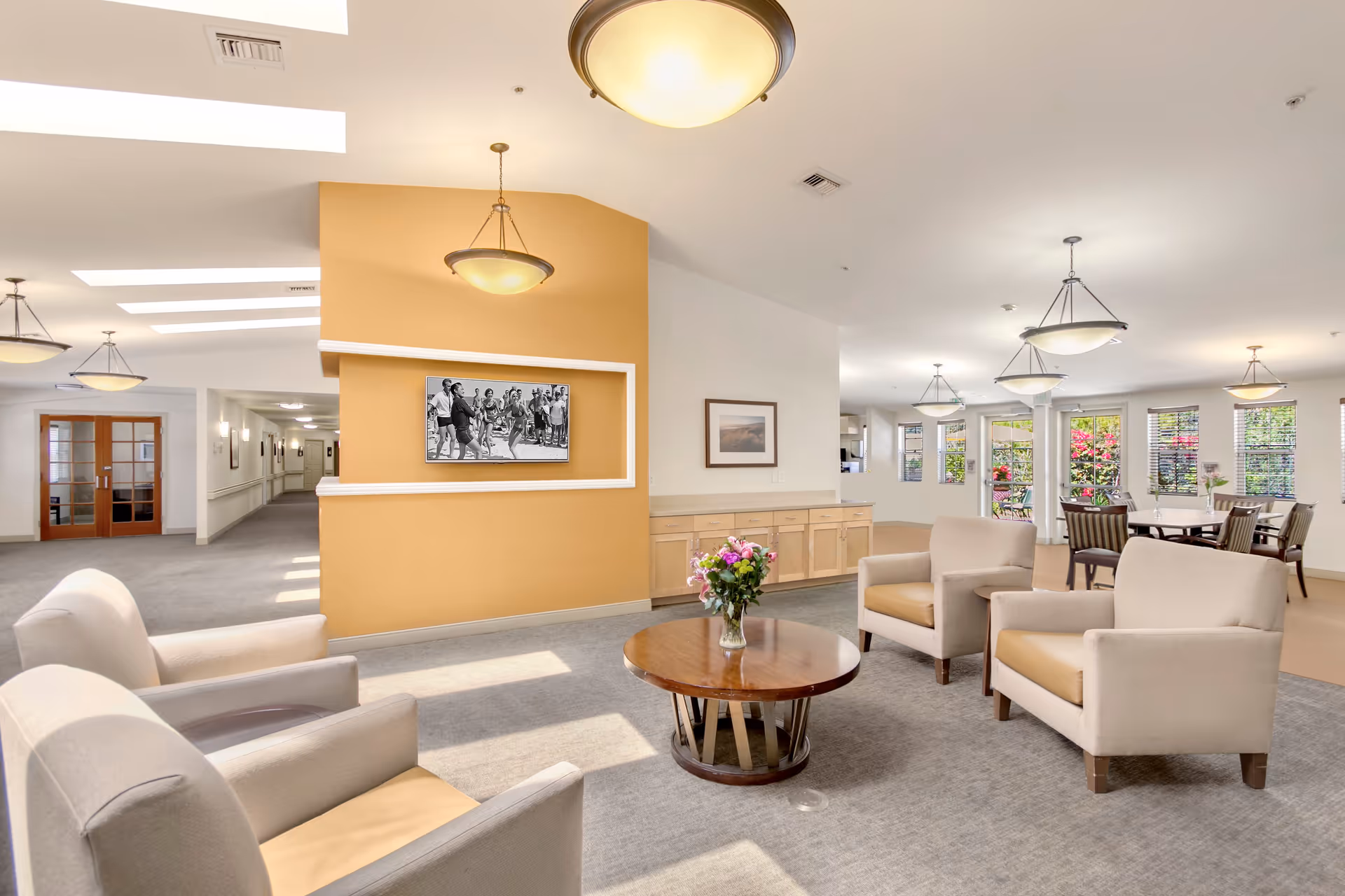 A bright and spacious common area in a senior living facility featuring four beige armchairs arranged around a round wooden coffee table with a vase of flowers. The room has large windows with blinds, multiple ceiling light fixtures, and a wall with a mounted black and white photograph. In the background, there are dining tables and chairs near the windows, and a hallway with double wooden doors.