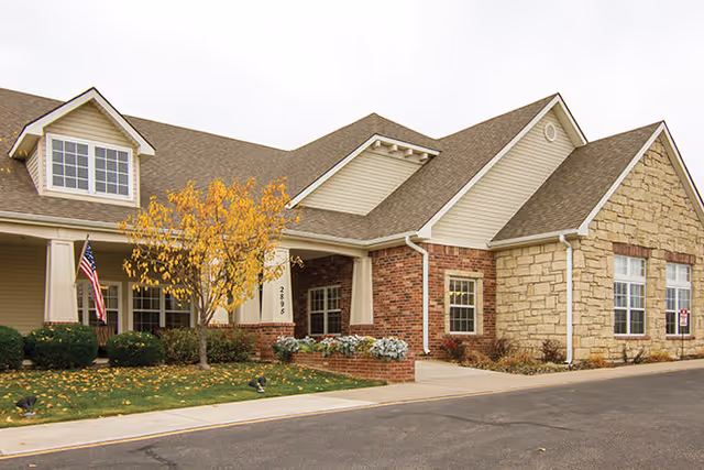 Exterior view of a single-story building with a combination of brick, stone, and siding. The building has a gabled roof with dormer windows, a small porch with white columns, an American flag, and a tree with yellow autumn leaves in front. The address number 2895 is visible near the porch.