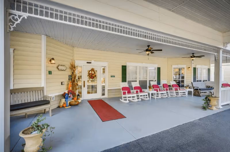 Covered porch area at the entrance of The Legacy of Hartsville facility featuring a row of white rocking chairs with red cushions, ceiling fans, benches, potted plants, and seasonal decorations near the door.
