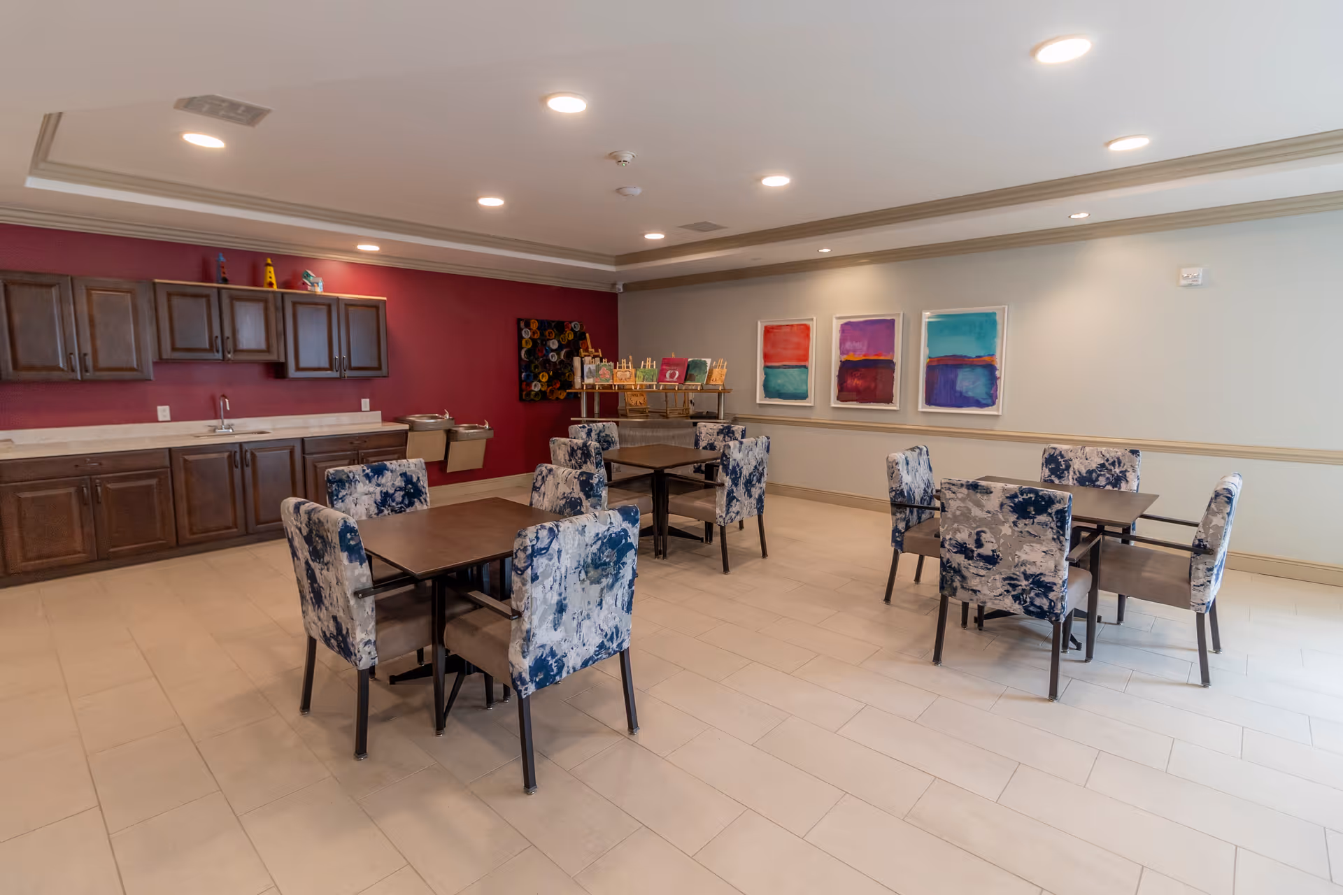 A dining area with four square tables, each surrounded by four chairs with blue and white patterned upholstery. The room has a tiled floor, a maroon accent wall with dark wooden cabinets and a sink, and three colorful abstract paintings on a light-colored wall. There is also a water fountain and a small shelf with decorative items.