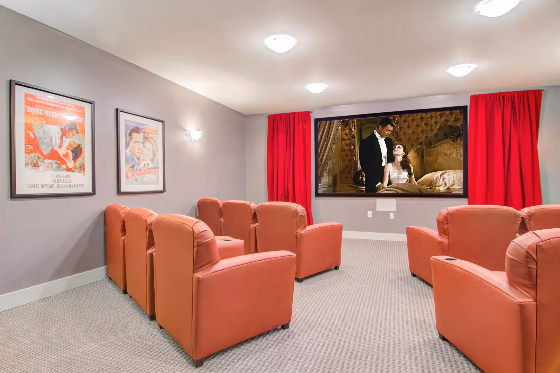 Small screening room with rows of red upholstered recliner chairs facing a large wall-mounted movie screen framed by red curtains.