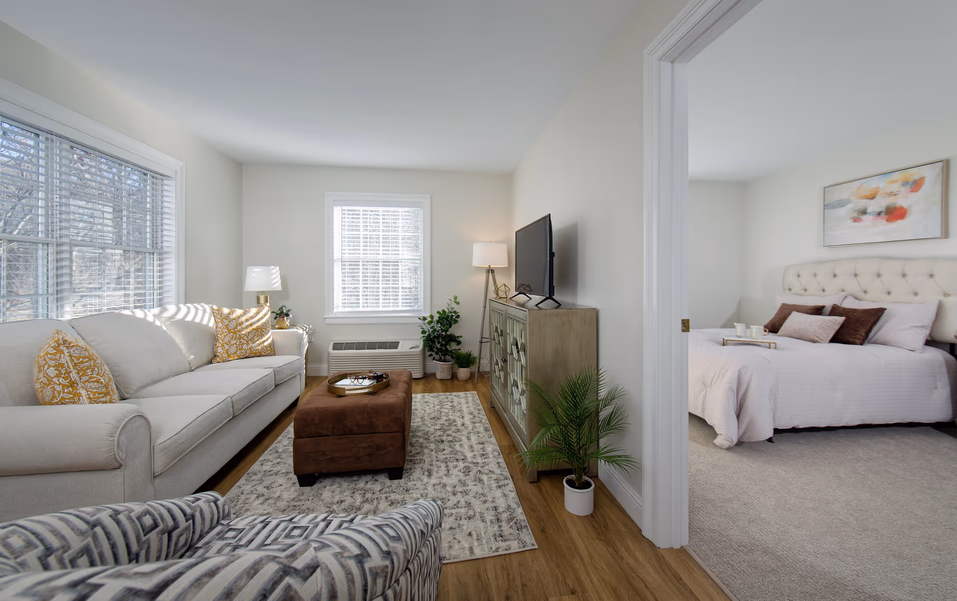 A bright living room with a beige sofa adorned with yellow patterned pillows, a brown ottoman, a patterned armchair, a TV on a wooden cabinet, and several potted plants. Adjacent to the living room is a bedroom with a tufted headboard bed, light pink bedding, and a tray with cups on the bed. The room has large windows with white blinds allowing natural light to fill the space.
