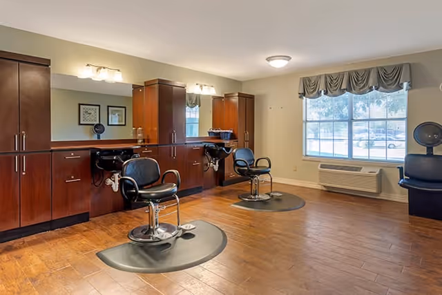 Interior view of a hair salon area in a senior living facility with two black salon chairs in front of wooden cabinets and mirrors. The room has wooden flooring, a large window with a valance, and a wall-mounted air conditioning unit below the window.