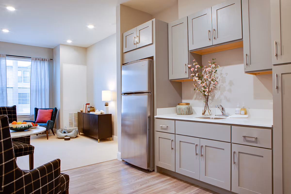 A modern kitchen area with light gray cabinets, a stainless steel refrigerator, and a countertop with a sink. A vase with pink flowers and soap dispensers are on the counter. Adjacent to the kitchen is a living area with a window covered by light curtains, a dark armchair with a red pillow, a round table with a bowl of fruit, and a dark sideboard with a lamp and decorative items.