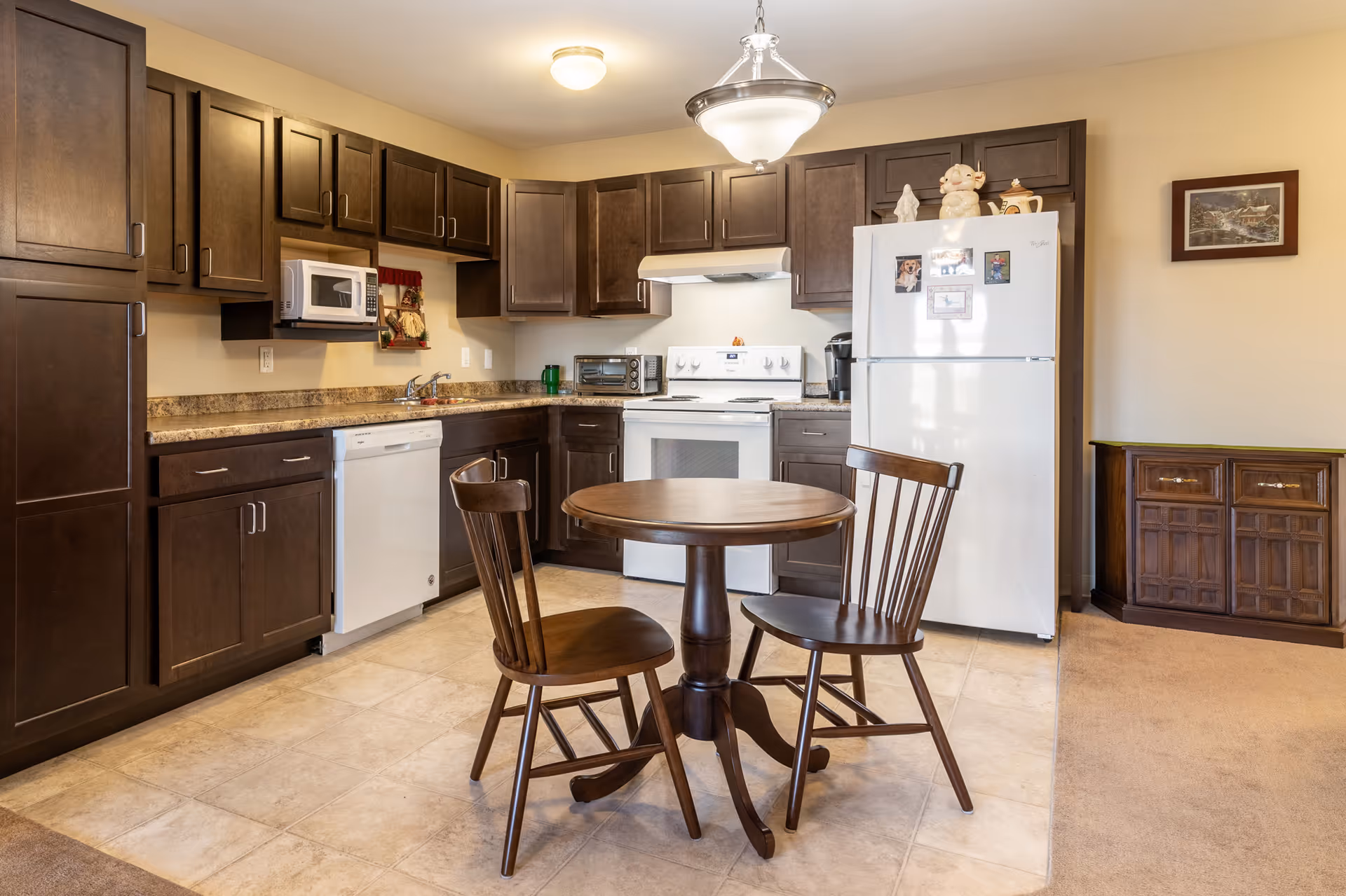 Small kitchen with dark wood cabinets, white appliances, and a round wooden table with two chairs.