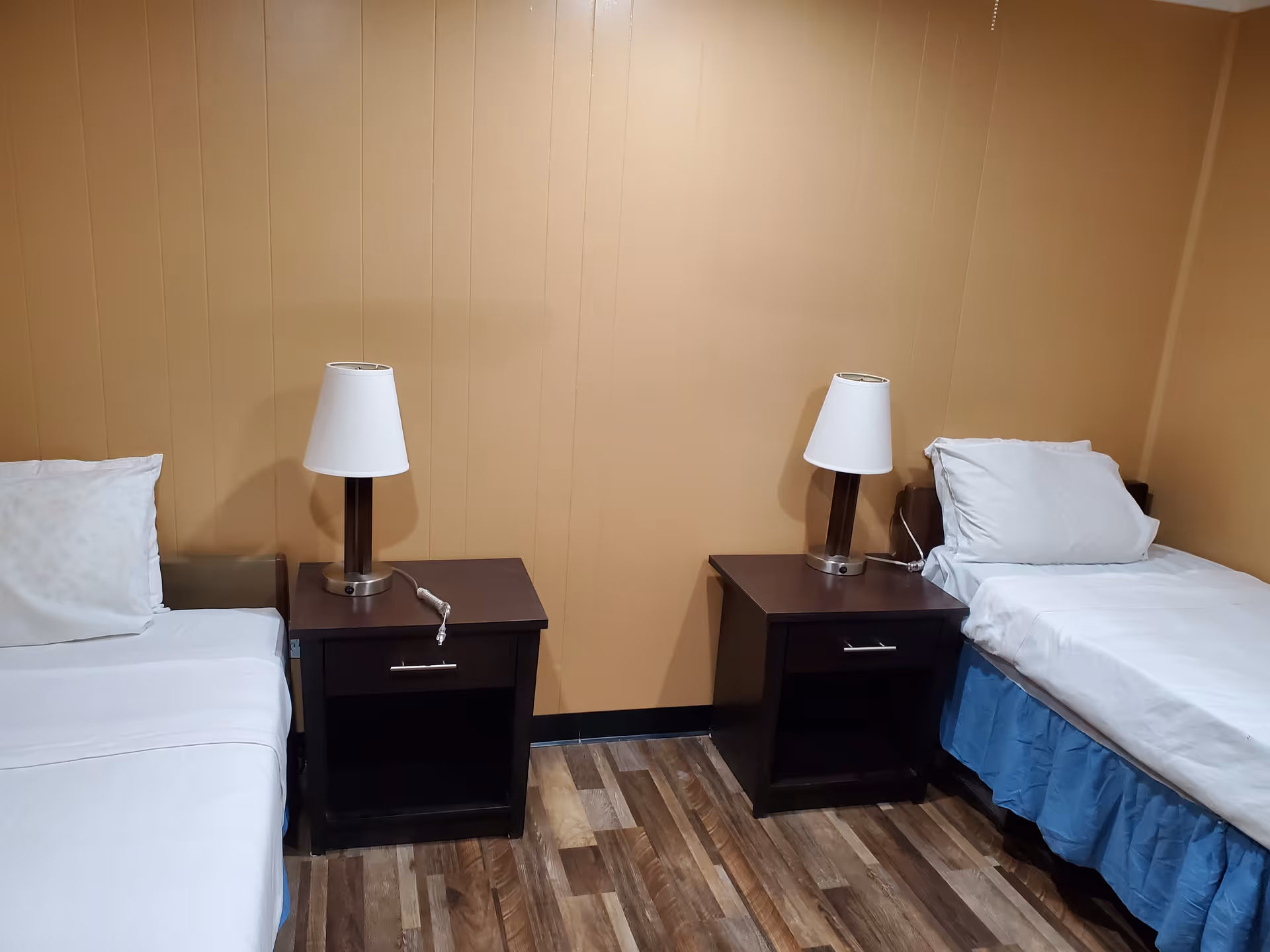 A small bedroom with two single beds separated by two dark wooden nightstands, each with a white lamp. The beds have white sheets and pillows, and the floor is wood-patterned. The walls are painted a light brown color.