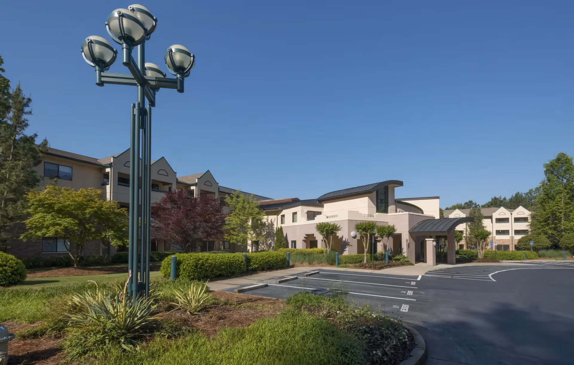 Exterior view of Delmar Gardens of Gwinnett Senior Living facility showing a multi-story building with beige and light brown walls, surrounded by landscaped greenery including trees and bushes, under a clear blue sky. There is a parking lot in front with marked parking spaces and a tall street lamp with multiple round lights.
