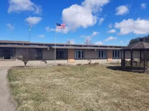 Single-story senior living facility building with a brick and beige exterior, an American flag on a flagpole in front, a gazebo to the right, and a grassy lawn under a partly cloudy blue sky.