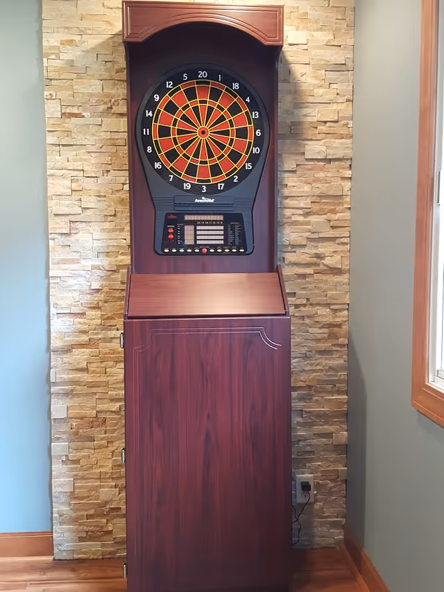 Electronic dartboard mounted on a wooden cabinet against a stone accent wall in a room with light blue walls and a window with wooden trim.