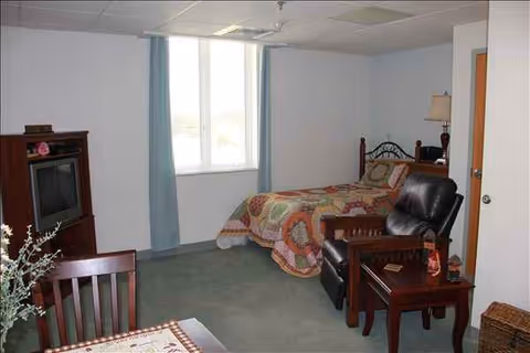 A furnished bedroom in a senior living facility with bed, seating, and a TV.