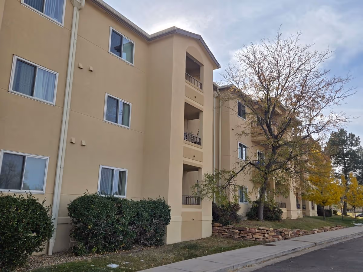Beige three-story retirement building with balconies, windows, shrubs and a sidewalk lined by trees.