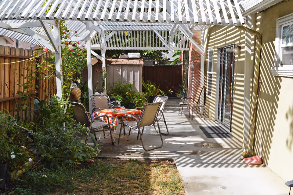 Outdoor patio area with a white pergola casting striped shadows on the ground and walls. There is a table covered with a colorful polka dot tablecloth surrounded by four chairs. The patio is adjacent to a yellow building with a sliding glass door and a window. There are plants and flowers along a wooden fence on the left side and a small shed in the background.