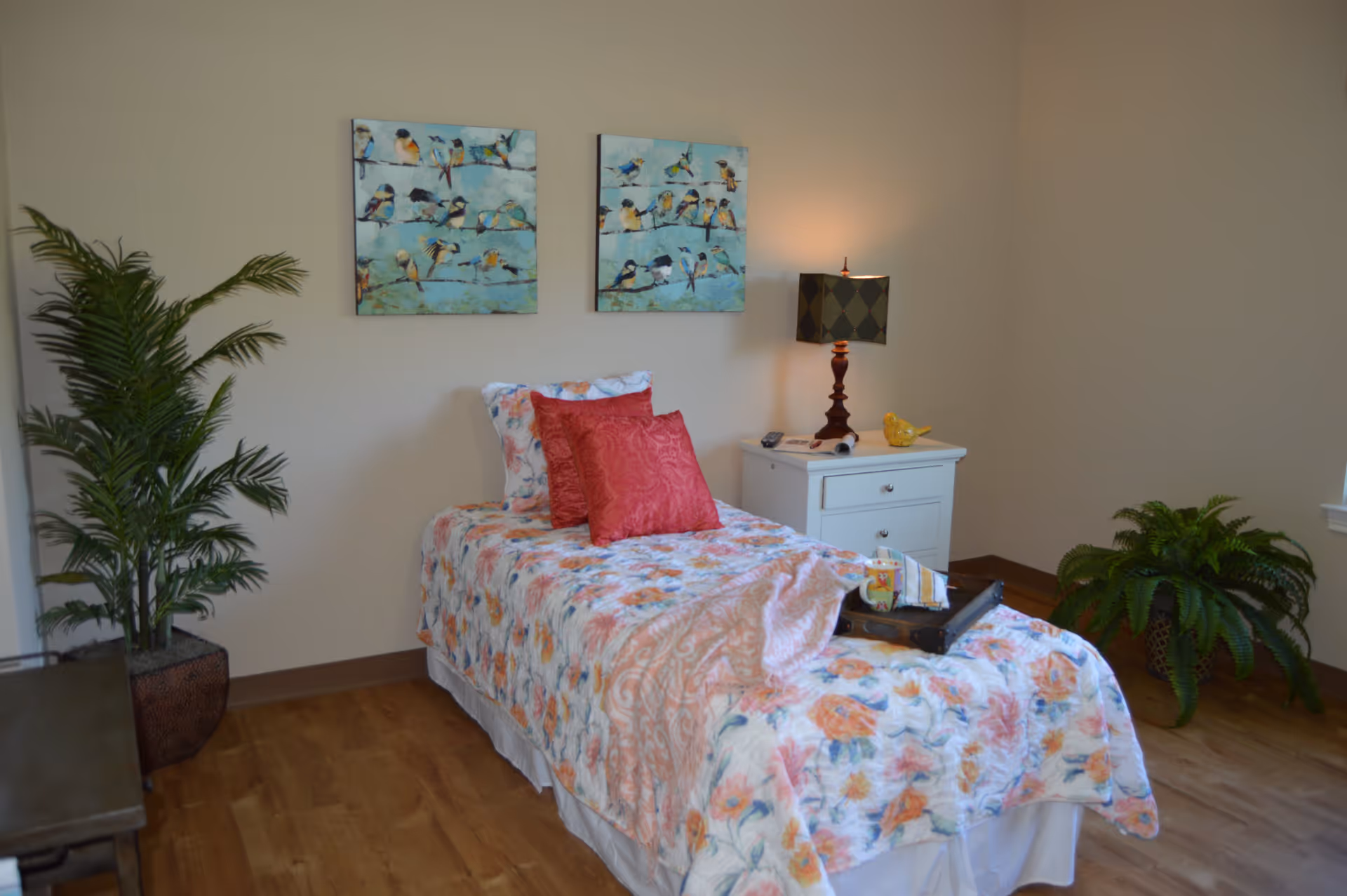 A cozy bedroom with a single bed covered in a floral bedspread and two red pillows. There is a white nightstand next to the bed with a lamp, a small yellow bird decoration, and a remote control. Two paintings of birds on branches hang on the wall above the bed. The room has wooden flooring and two green potted plants, one on the left side and one near the window on the right.