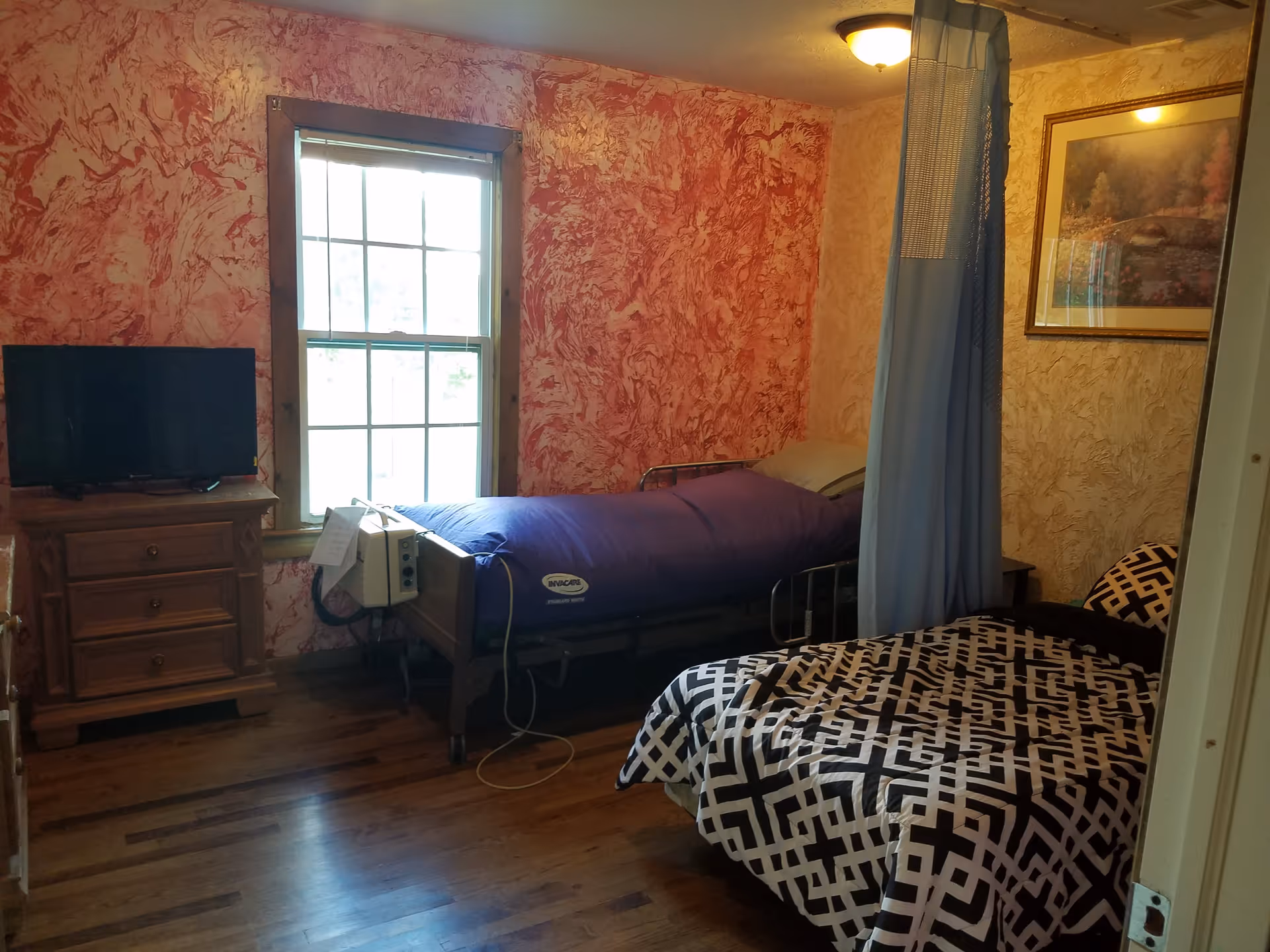 A room with two beds, one hospital-style bed with a purple cover and medical equipment attached, and another bed with a black and white geometric patterned bedspread. The room has wooden floors, a window with natural light, a small wooden dresser with a TV on top, a blue privacy curtain, and a framed picture on the wall.