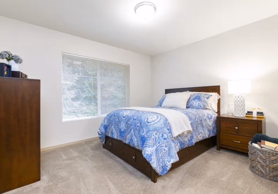 Bright bedroom with a wooden bed dressed in blue patterned bedding, a nightstand with a lamp, dresser, and a window with blinds.