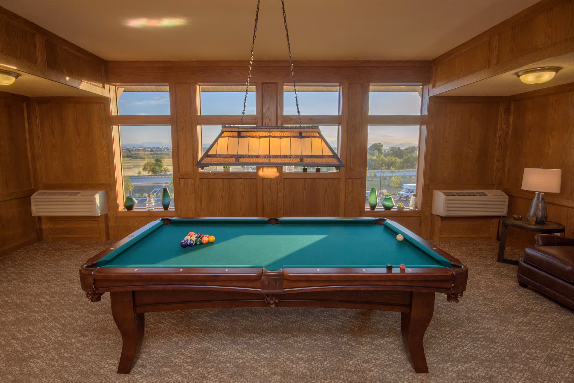 A cozy indoor room with a green felt pool table in the center, set up for a game with billiard balls arranged in a triangle and a cue ball nearby. The room features wood-paneled walls, large windows letting in natural light, two air conditioning units, a hanging light fixture above the pool table, and a leather armchair with a side table and lamp in the corner.