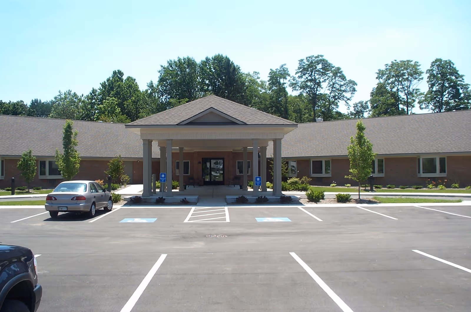 Front exterior view of Greenleaf Health Campus showing a single-story building with a covered entrance supported by columns, surrounded by a parking lot with marked handicap spaces and some small trees and shrubs.