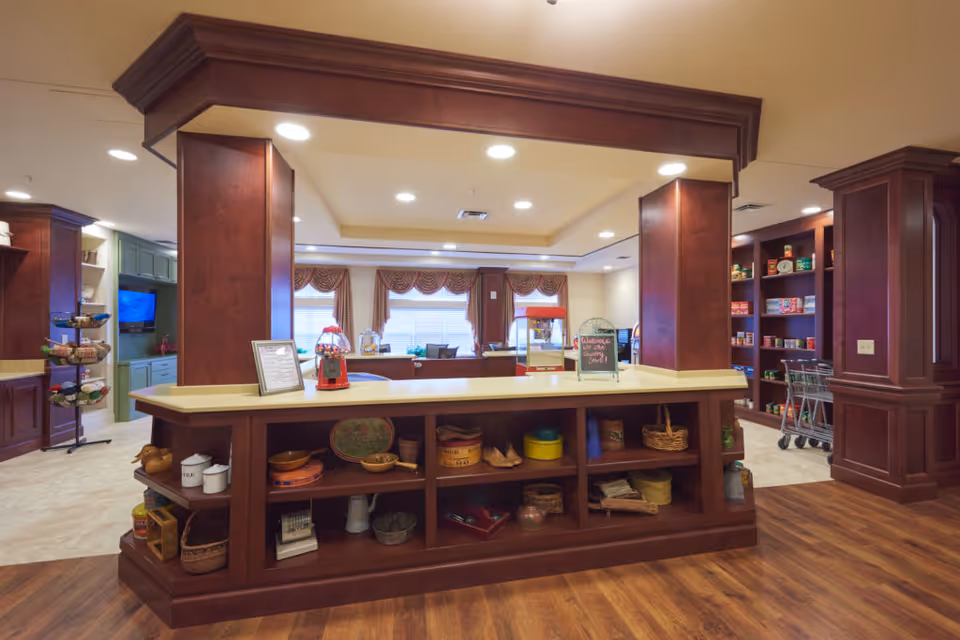 Interior view of a senior living facility's common area with a large wooden counter in the center displaying various decorative items. Behind the counter, there are windows with curtains, a popcorn machine, and shelves stocked with canned goods and other items. The room has warm lighting and wooden flooring.