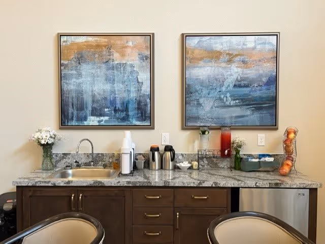 A kitchenette area with a granite countertop, a small sink, two abstract paintings on the wall, a vase with white flowers, coffee dispensers, cups, a container with creamers, a red beverage dispenser, and a basket with apples. Two chairs with beige cushions are partially visible in the foreground.