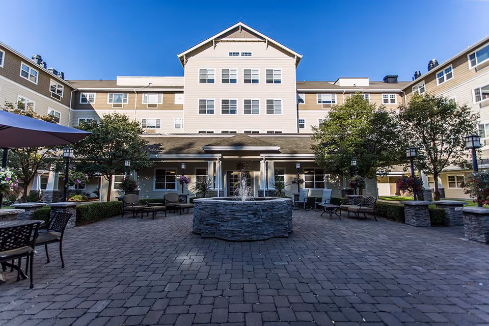 Outdoor courtyard area of Garden Way Retirement Community featuring a central stone fountain, surrounded by patio seating with chairs and tables, trees, and hanging flower baskets, with the multi-story building in the background under a clear blue sky.