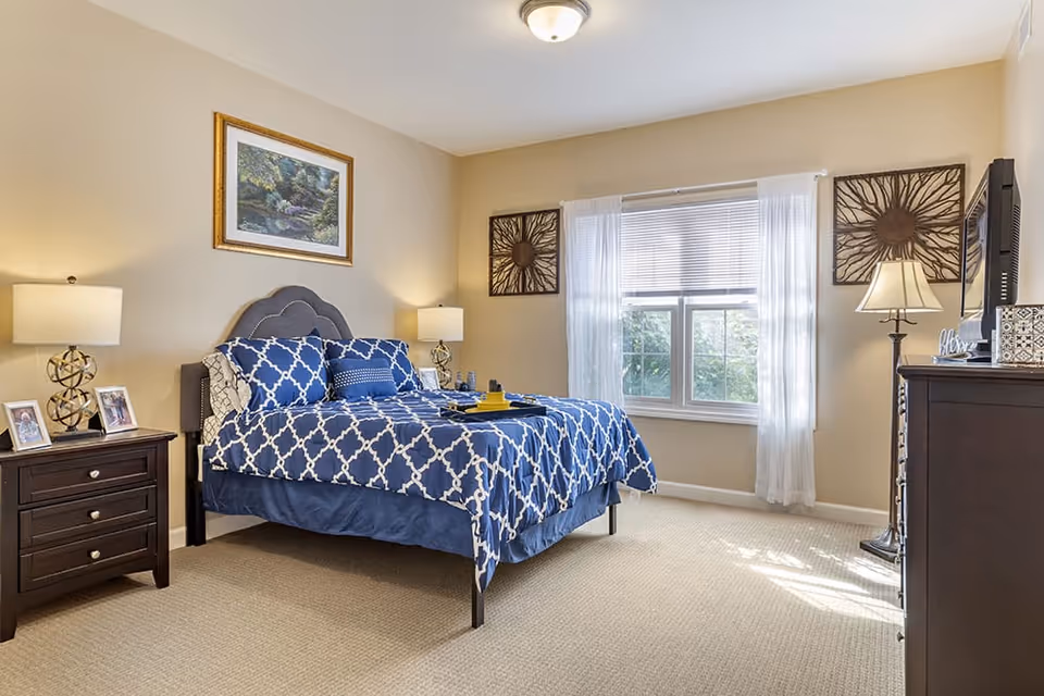 A well-lit bedroom with a bed dressed in blue and white patterned bedding. The room features beige walls, a window with white curtains, two bedside tables with lamps, framed artwork on the walls, and a dark wood dresser with a TV on top.