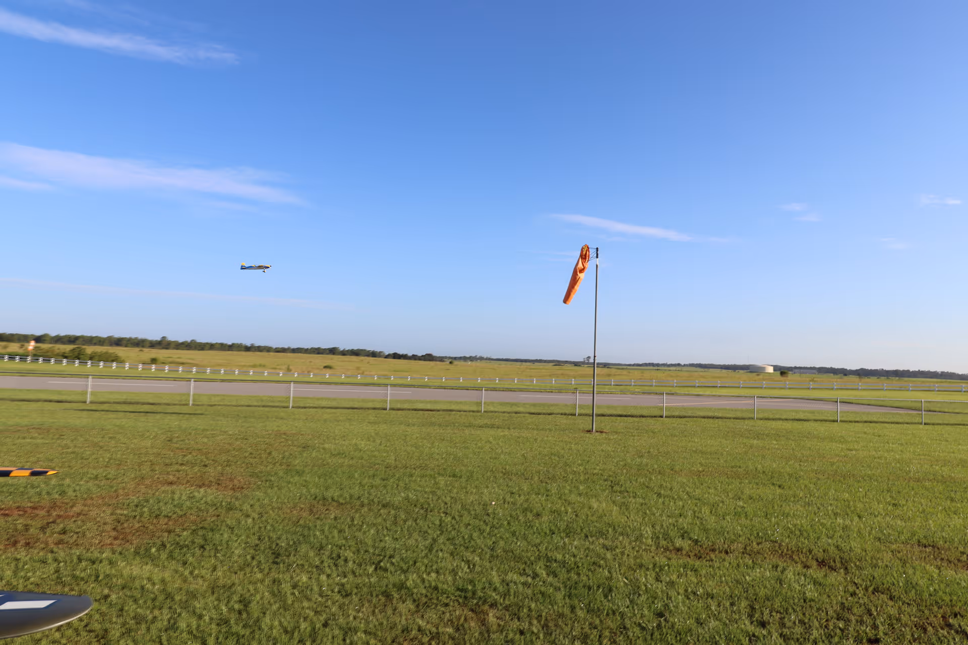 Grassy airfield with a windsock and a small airplane flying under a clear blue sky.