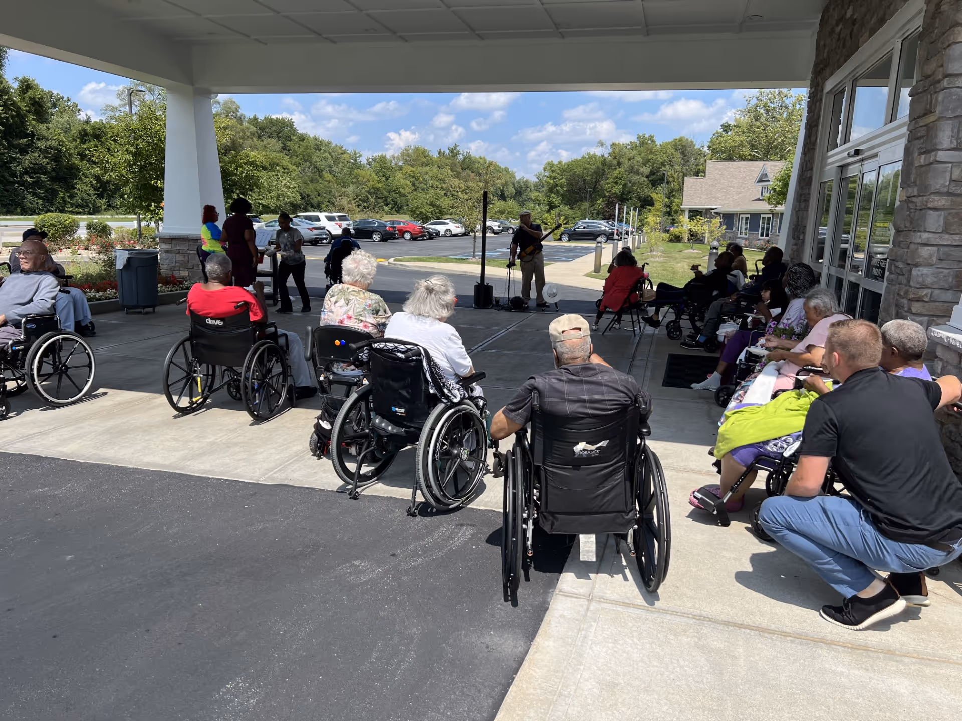 A group of elderly people, many in wheelchairs, gathered outside under a covered area at Creekside Health and Rehabilitation Center. They are seated facing a musician playing a guitar and singing. The setting is sunny with a parking lot and trees in the background.