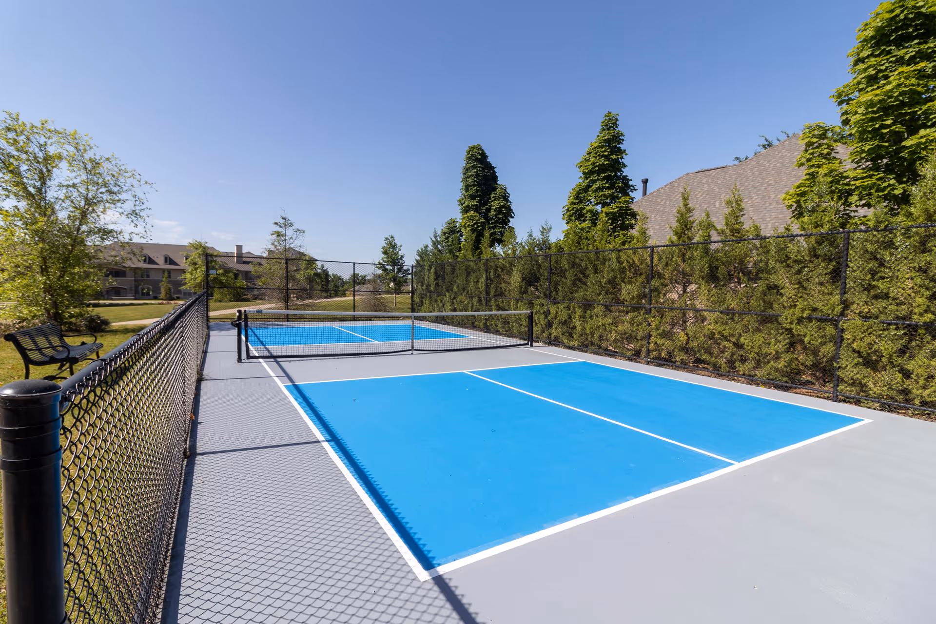 Two outdoor blue pickleball courts surrounded by a chain-link fence, benches, and trees under a clear blue sky.