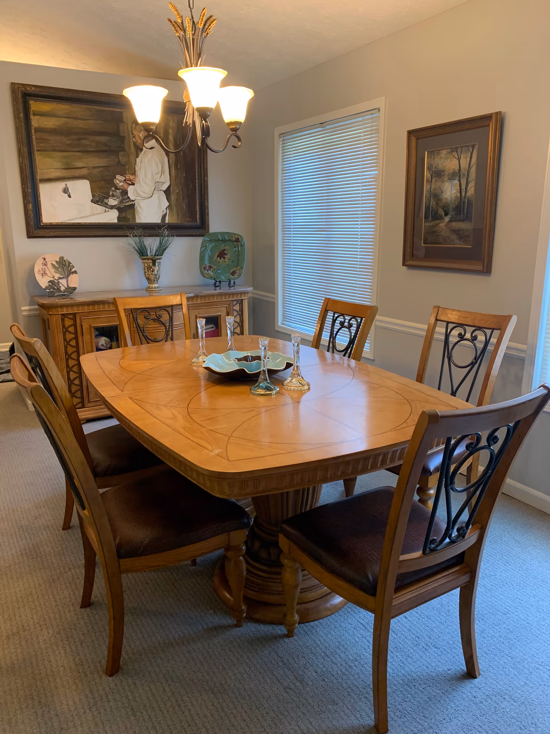 Wooden dining table with six chairs under a chandelier, a sideboard, and framed artwork in a dining room.