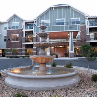 A multi-tiered stone fountain in front of the entrance to a multi-story senior living building with balconies and decorative siding.