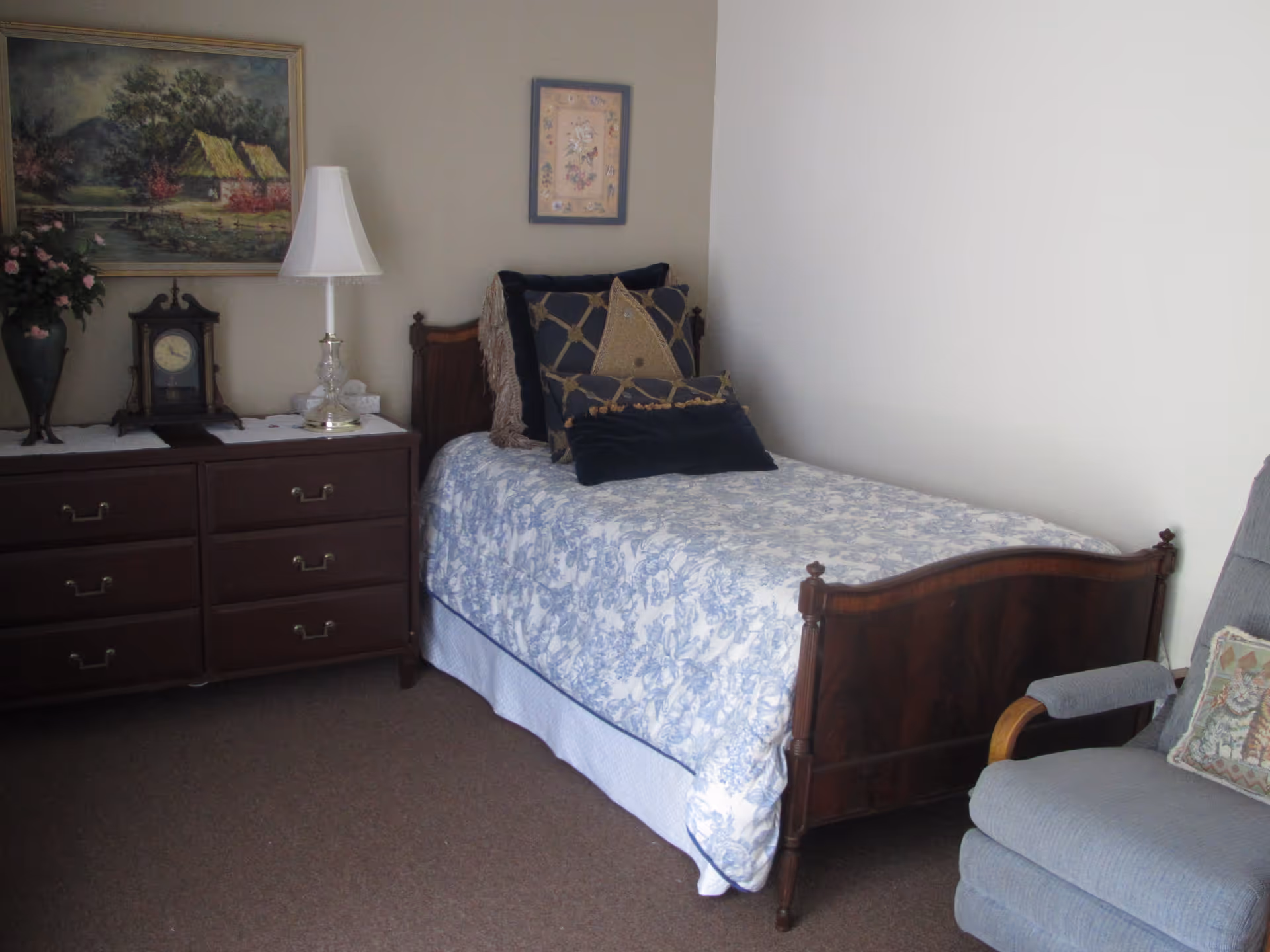 A bedroom with a single wooden bed covered with a blue and white floral bedspread and several decorative pillows. Next to the bed is a wooden dresser with a white lamp, a clock, and a vase with flowers. There are two framed pictures hanging on the walls. A blue upholstered armchair with a cat-themed pillow is positioned near the bed.