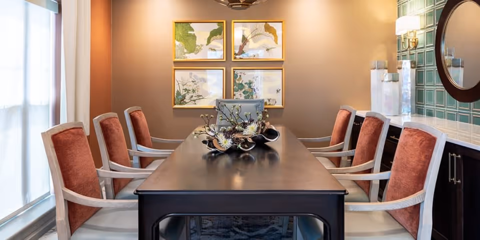 A dining room with a long dark wooden table surrounded by eight chairs with red cushions. A decorative centerpiece with flowers is placed on the table. The room has a large window with sheer curtains on the left, four framed botanical prints on the far wall, and a round mirror on the right wall above a cabinet with green tiled backsplash.