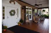 Covered entrance porch with a white door decorated with a wreath, rocking chairs, ceiling fans, potted plants, and brick exterior.