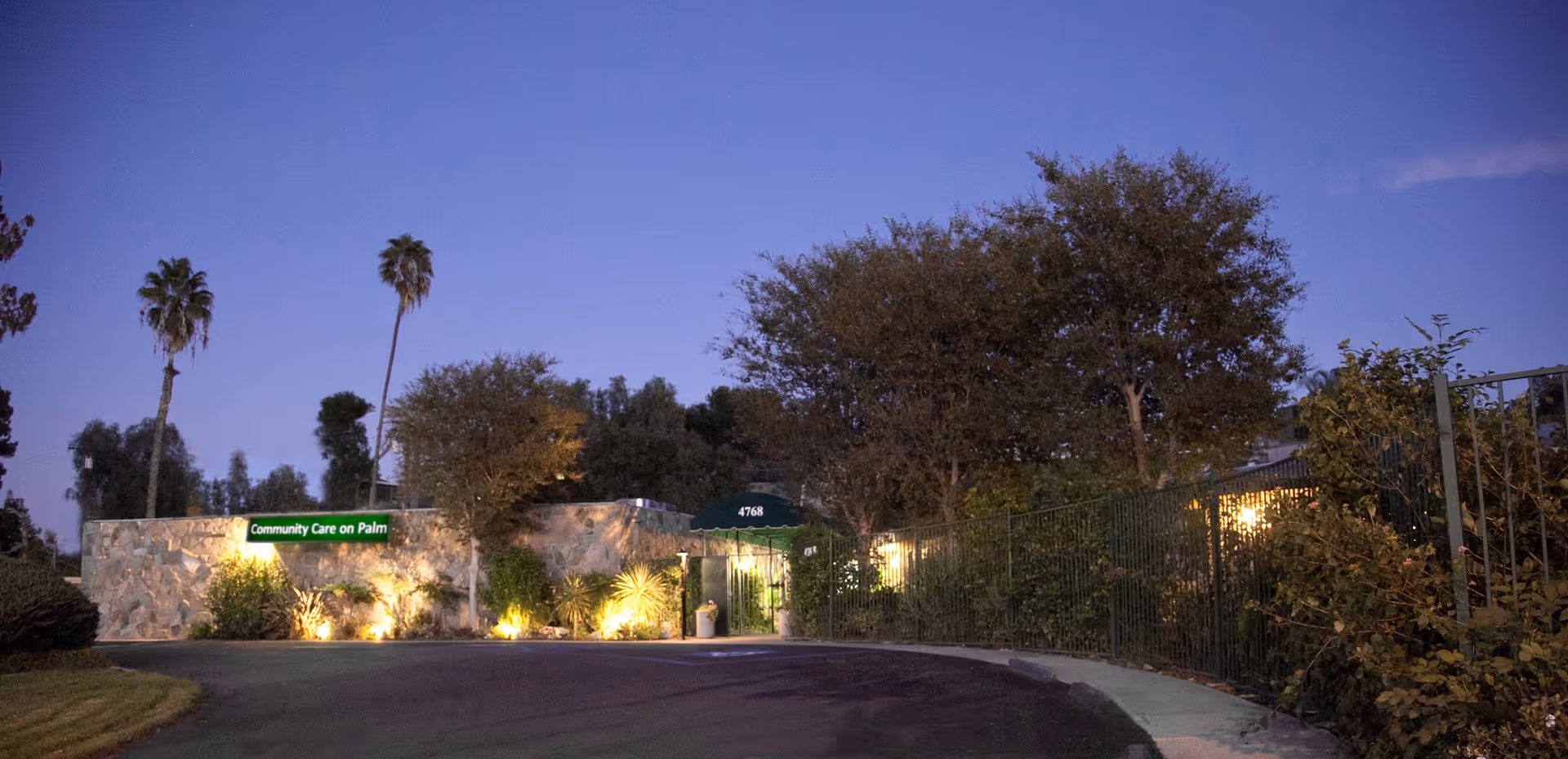 Exterior view of Community Care On Palm nursing facility at dusk, showing a stone wall with a green sign, trees, and a curved driveway leading to the entrance.