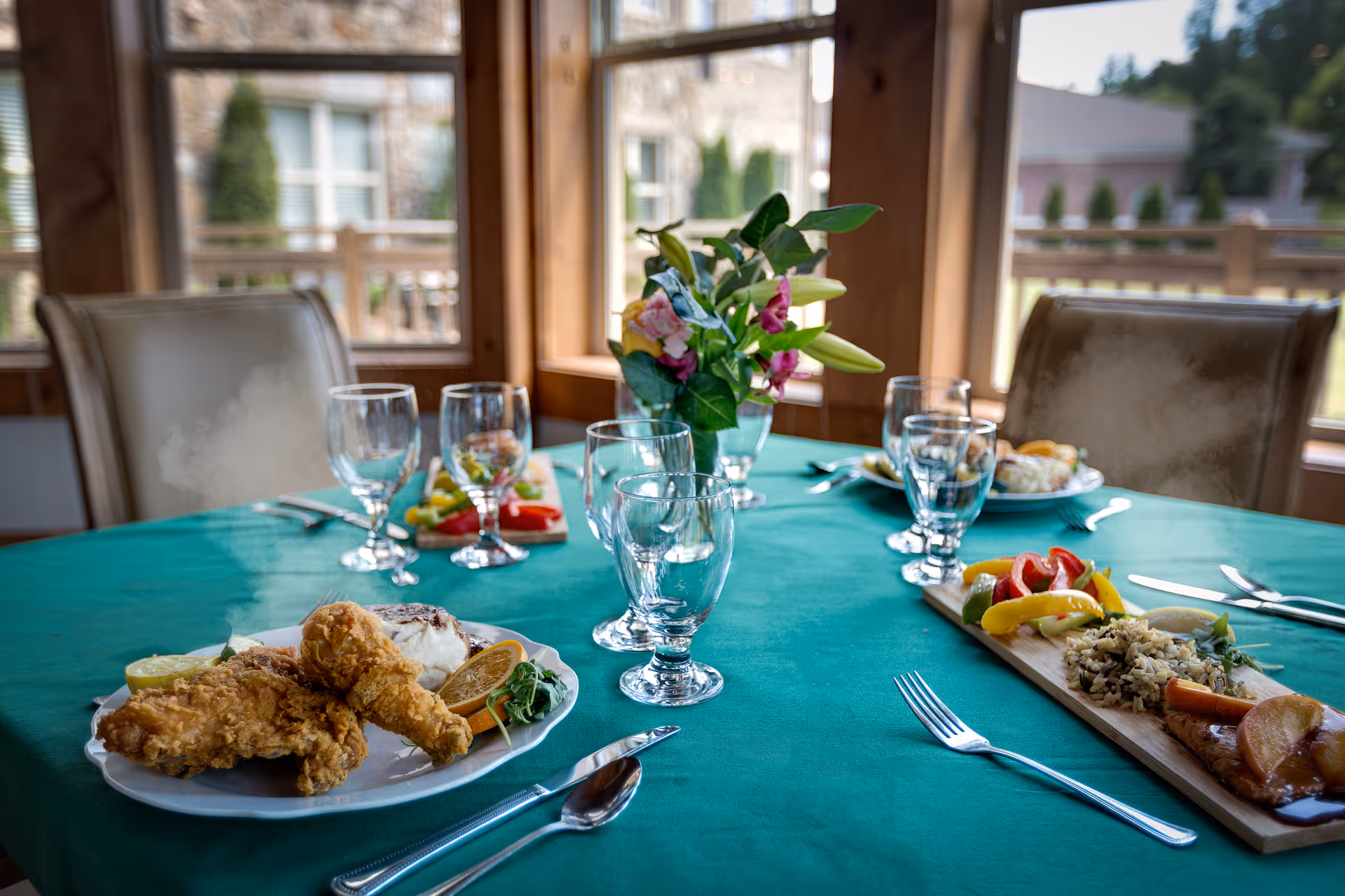 A dining table set with plates of food including fried chicken, rice with vegetables, and a colorful vegetable medley. There are multiple empty water glasses and silverware arranged on a green tablecloth. A small vase with flowers is placed in the center of the table. The background shows large windows with a view of an outdoor area and wooden chairs around the table.