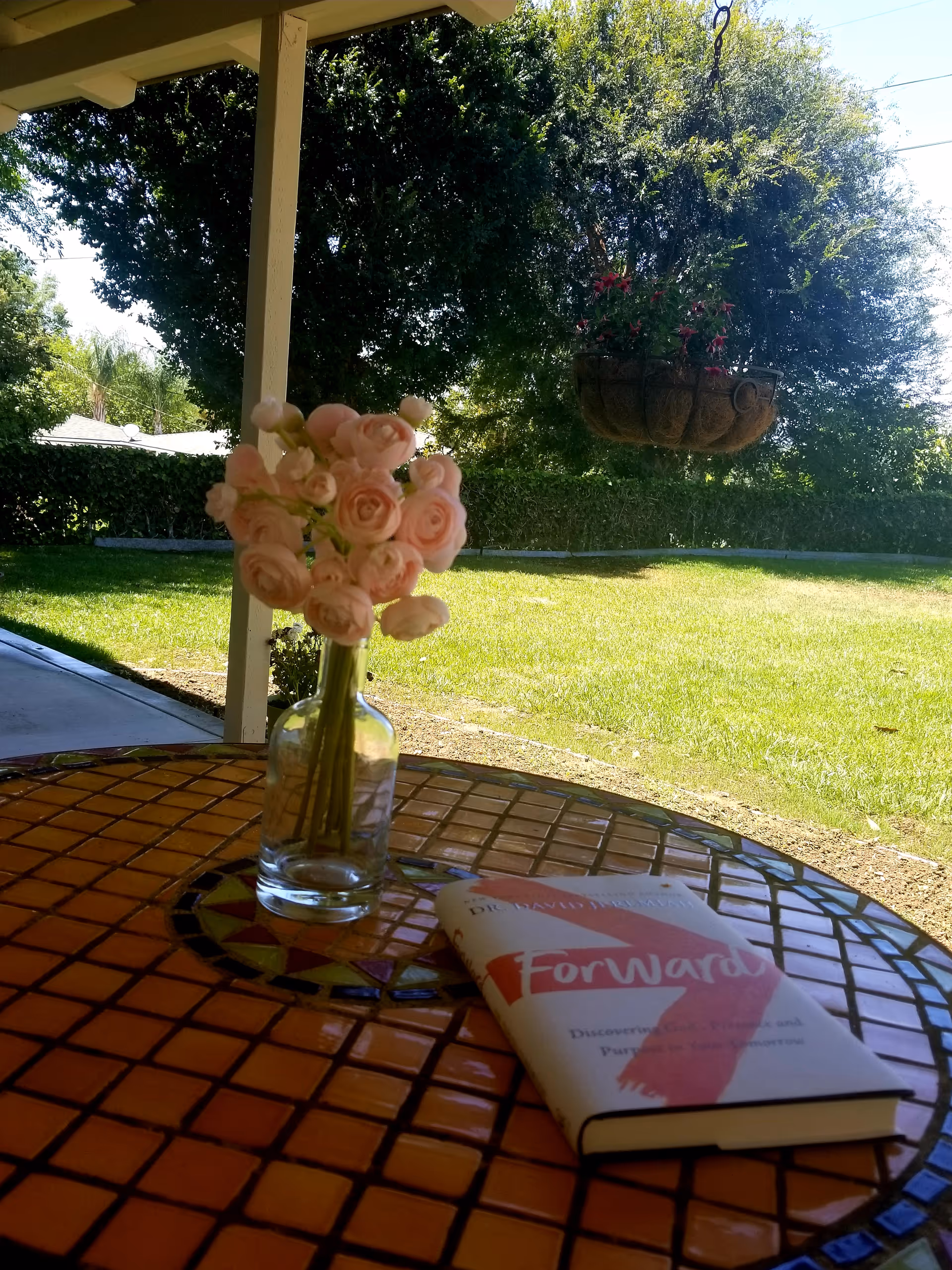 A round mosaic tile table with a glass vase holding light pink roses and a book titled 'Forward' placed on it. The table is on a covered patio overlooking a green lawn with trees and a hanging flower basket in the background.