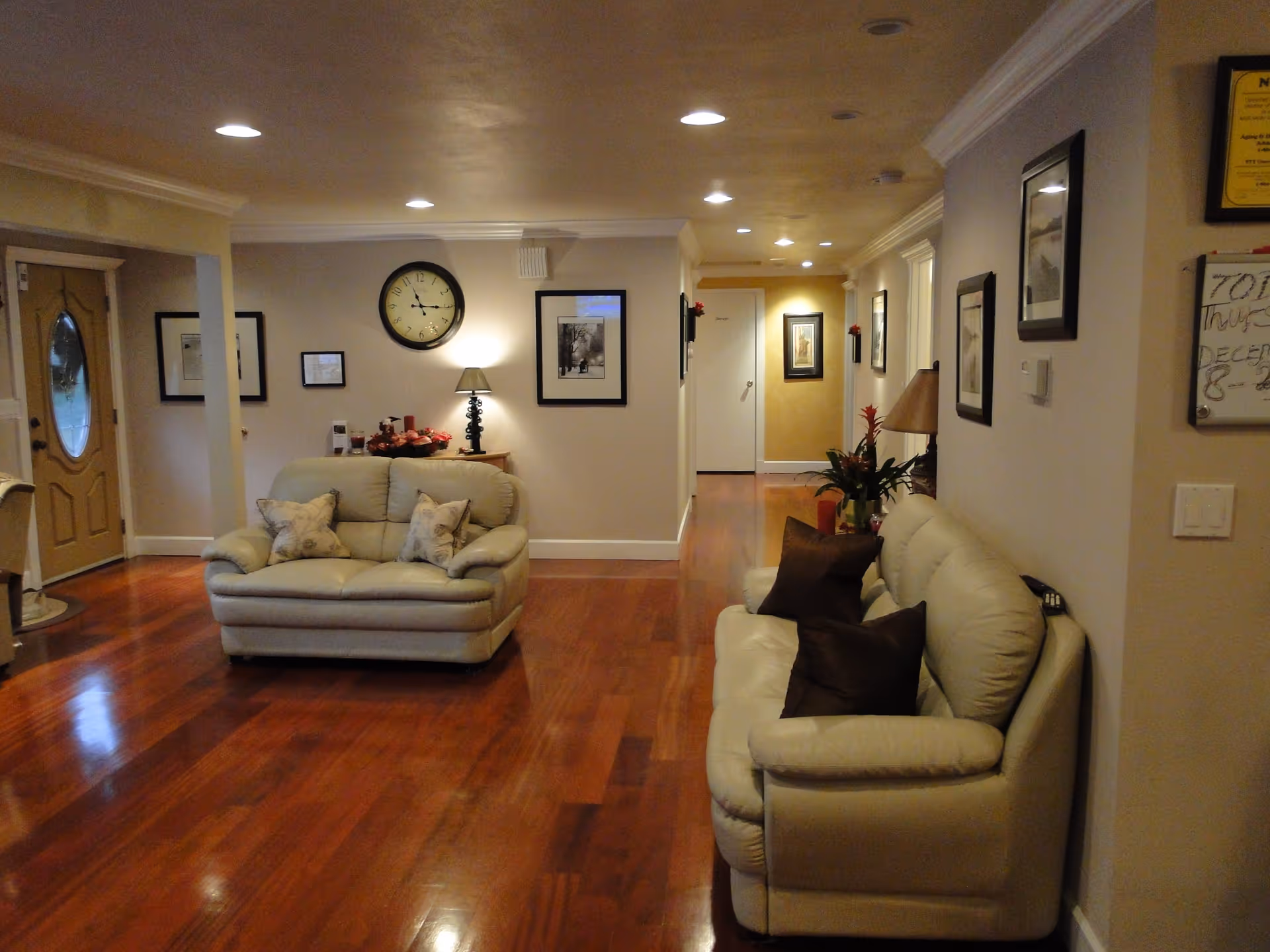 Interior view of a living room area with two cream-colored leather sofas, each with decorative pillows. The room has polished wooden floors, beige walls adorned with framed pictures, a round wall clock, and a table lamp on a small wooden table. There is a hallway leading to other rooms and a front door with an oval glass window on the left side.