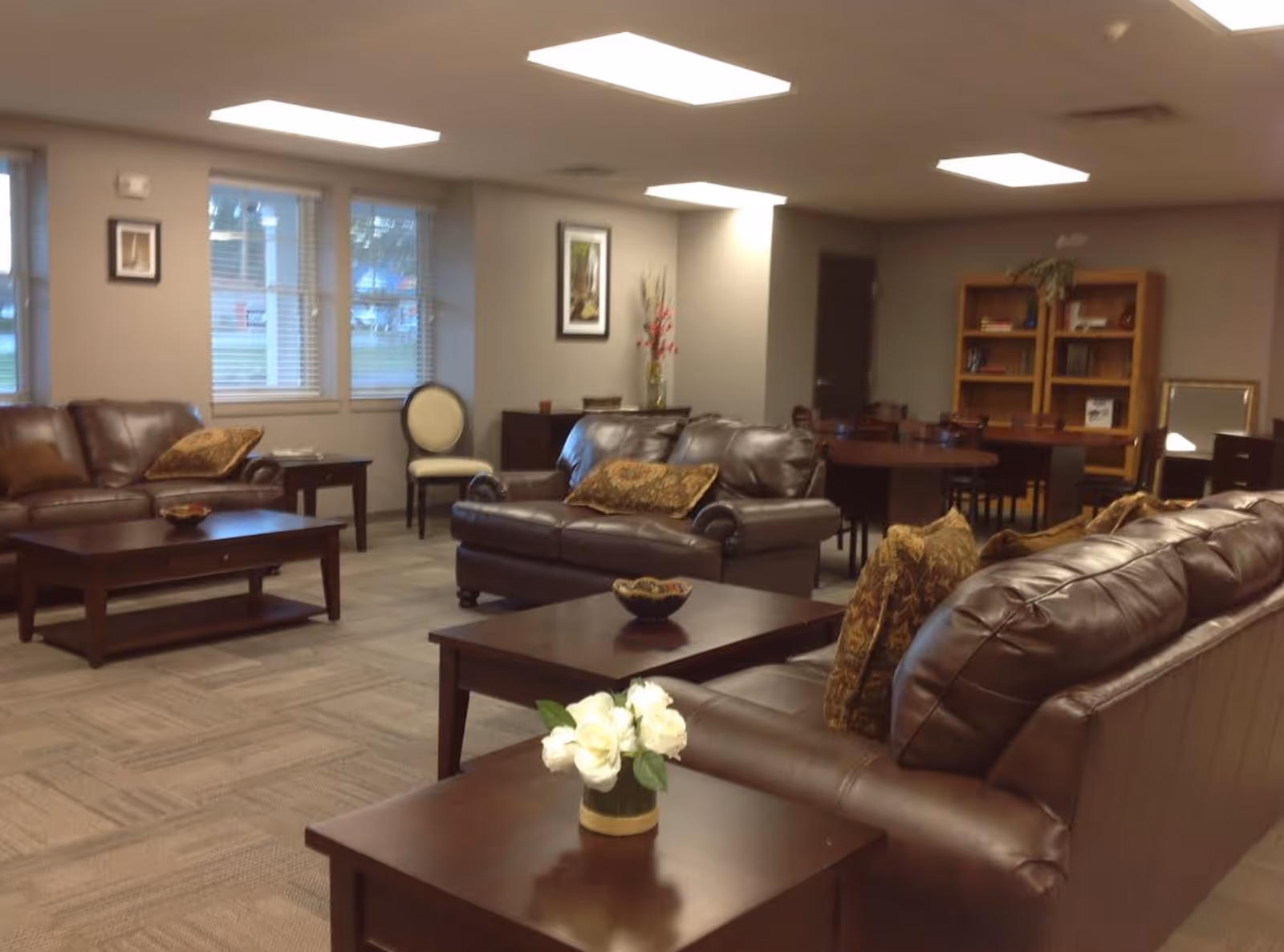 A cozy living room area in an assisted living facility with brown leather sofas, wooden coffee tables, a small vase with white flowers on one table, a bookshelf filled with books and decorative items, and a dining table with chairs in the background. The room has beige walls, carpeted floor, and windows with blinds.