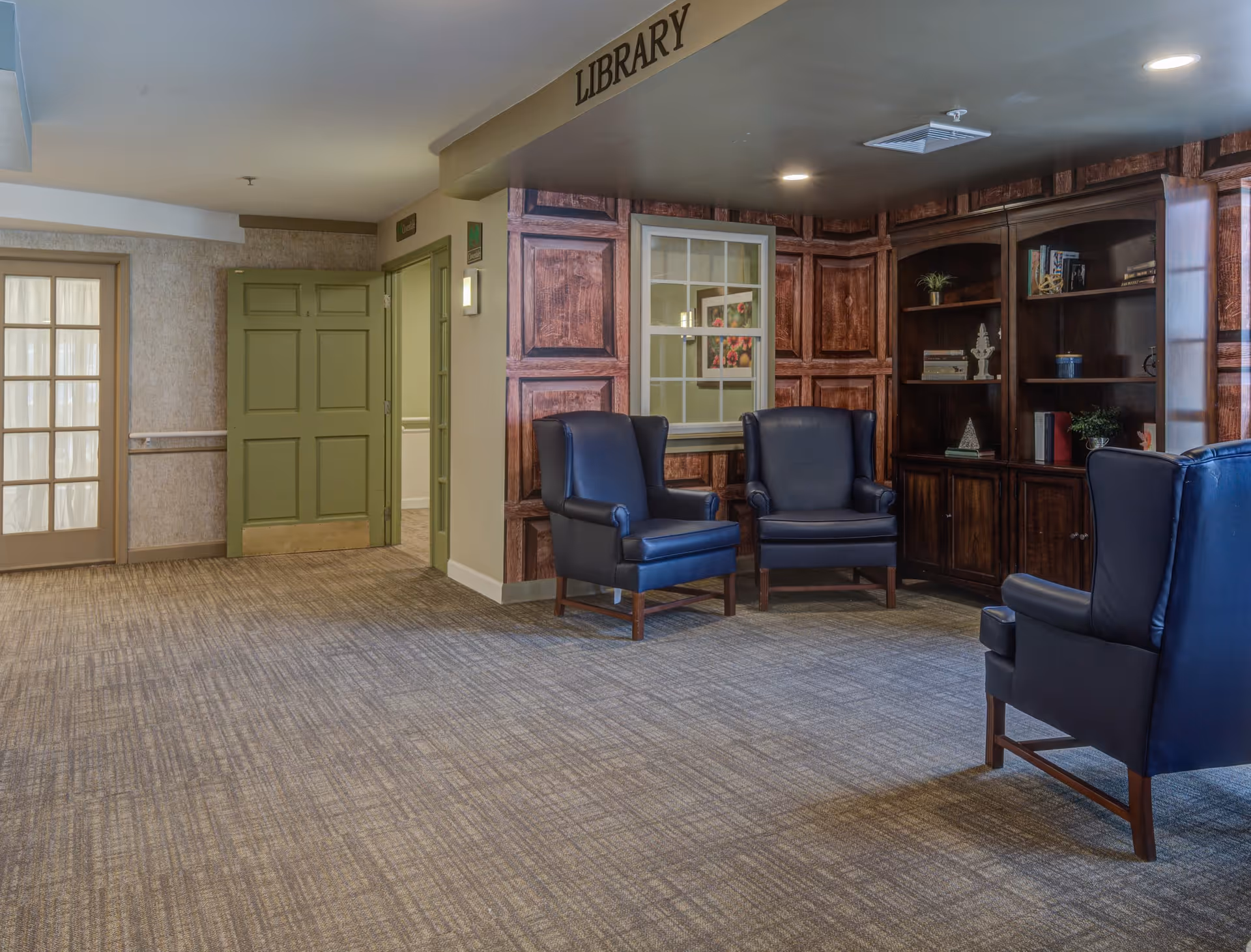 A quiet library area in a senior living facility with three dark blue armchairs arranged near a wooden bookshelf filled with books and decorative items. The walls have a wood panel design, and there is a sign above the area that reads 'LIBRARY'. The floor is carpeted, and there are green doors and a window with curtains in the background.
