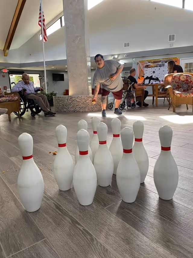An indoor scene at a senior living facility where a man is playing a game of indoor bowling with white pins set up on a wooden floor. Other residents are seated around the area, including a man in a wheelchair and two people sitting at a table in the background. The room has high ceilings with large windows allowing natural light to enter.