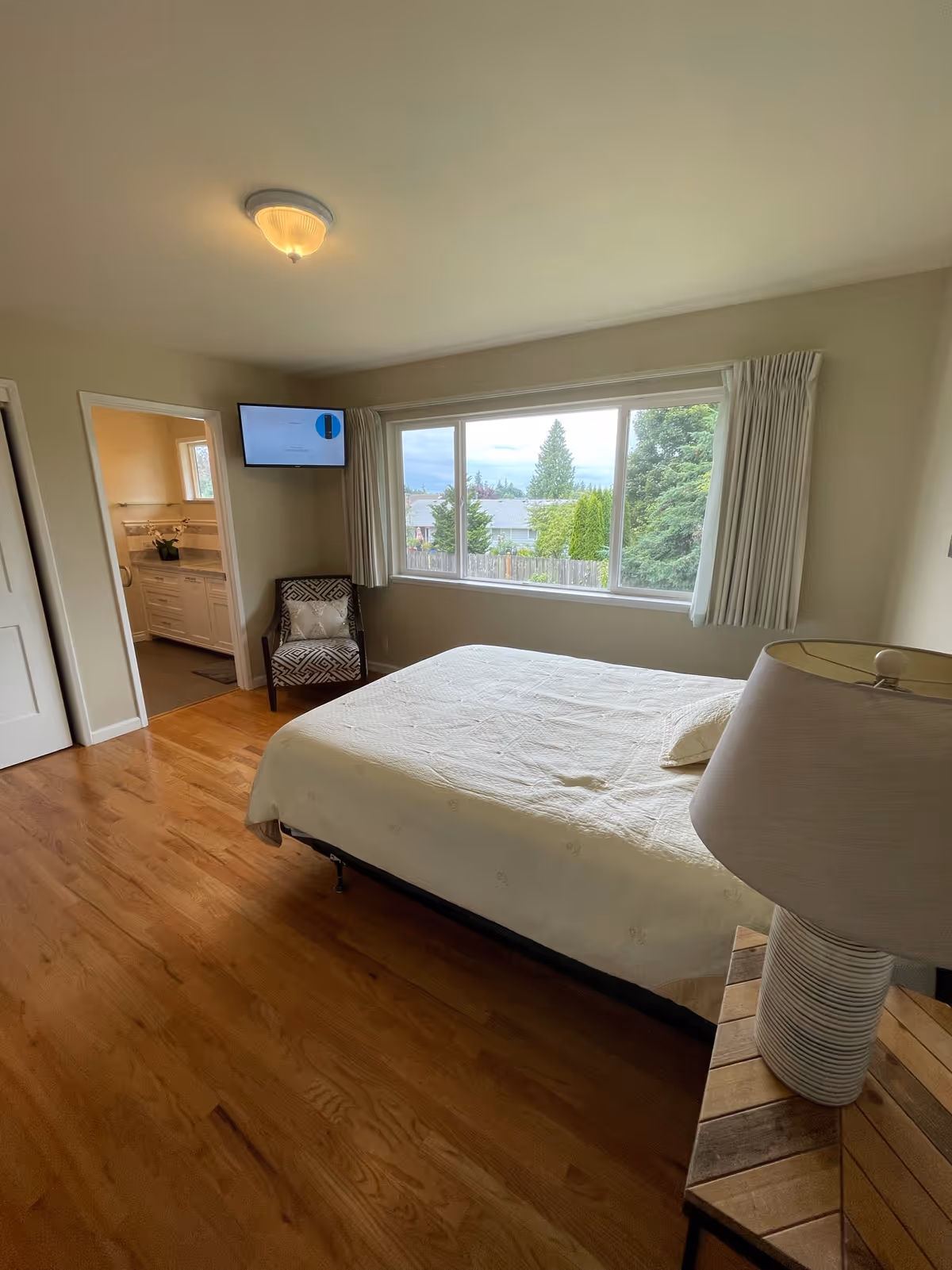 Bright bedroom with a bed, bedside lamp, armchair, wall-mounted TV, and a large window overlooking trees, with an open doorway to a bathroom.