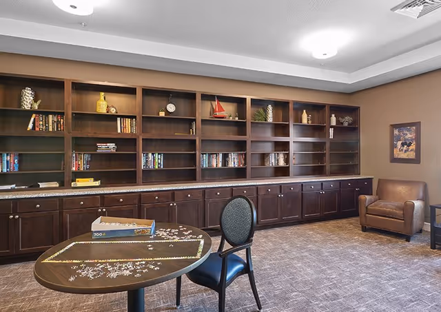 A senior living common room with a round table holding a jigsaw puzzle, chairs, built-in bookshelves and a leather armchair.