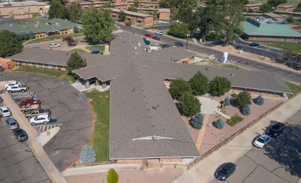 Aerial view of Rock Canyon Respiratory and Rehabilitation Center showing a large, single-story building with a cross-shaped roof, surrounded by trees, parking lots with several cars, and nearby streets.