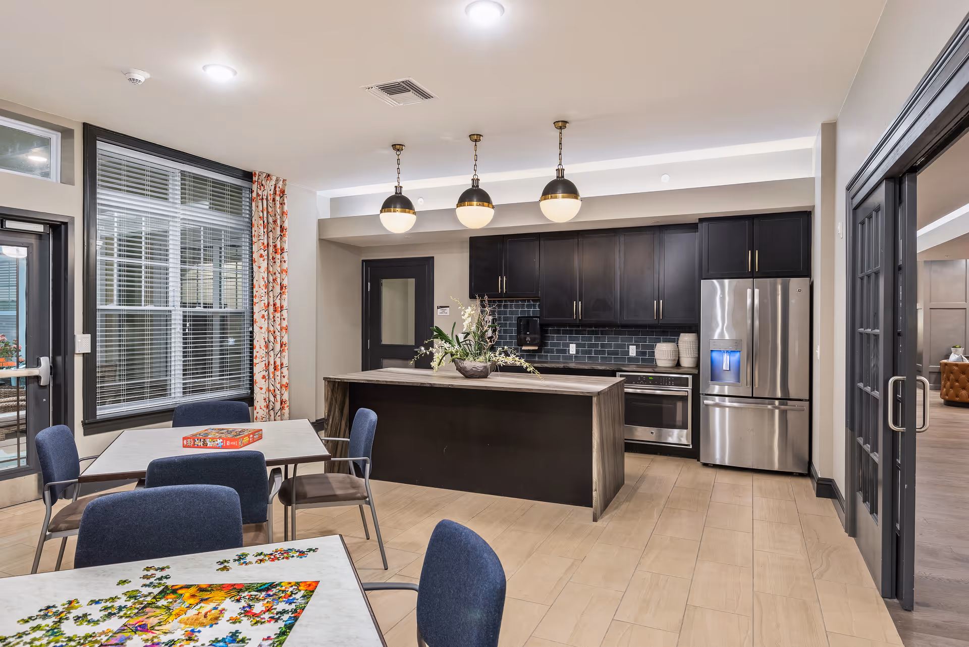 A modern kitchen and dining area in a senior living facility with a large island, dark cabinetry, stainless steel refrigerator, and three pendant lights. There are tables with chairs, one table has a partially completed jigsaw puzzle and another has a board game. Large windows with blinds and floral curtains let in natural light.