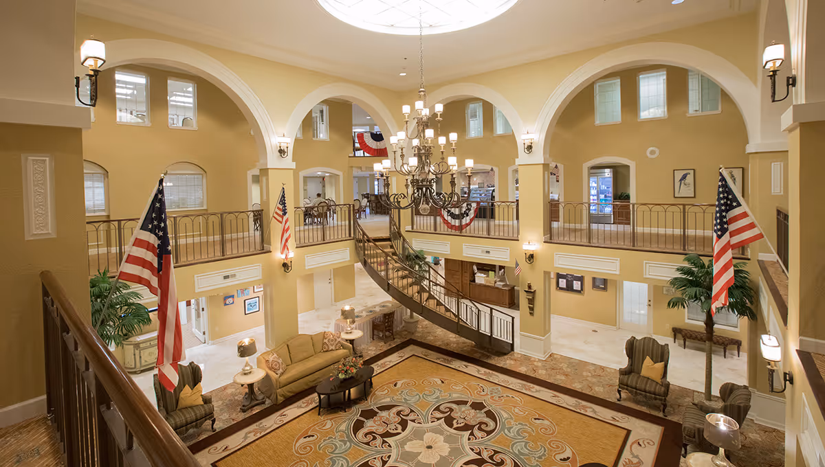 Spacious two-story lobby with a central chandelier, curved staircase, seating areas, arched balconies, and American flags.