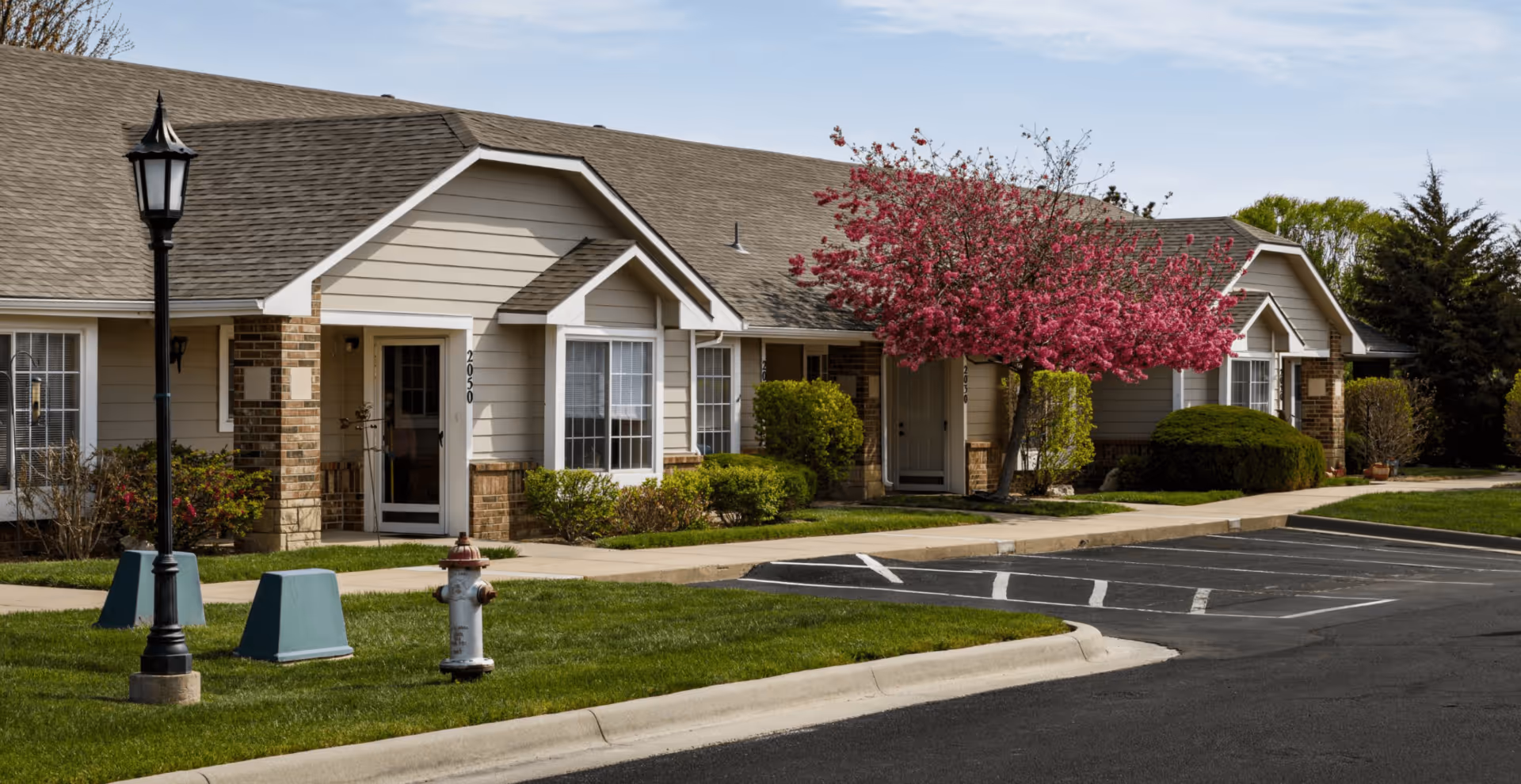 Single-story beige senior living cottages with a pink flowering tree, small front lawns, and an adjacent parking area.