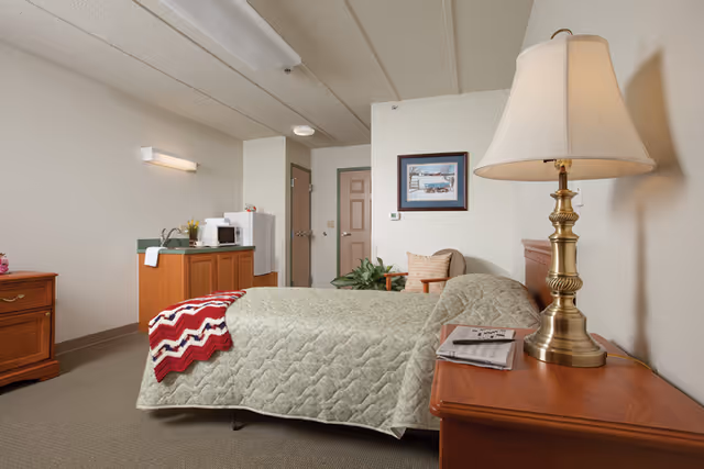 A senior living community bedroom featuring a single bed with a green quilt and a red and white knitted blanket at the foot. There is a wooden nightstand with a brass lamp and a newspaper on it. In the background, there is a small kitchenette area with a microwave, sink, and cabinets. A framed picture hangs on the wall above a cushioned chair with a pillow. The room has beige walls and carpeted floor.