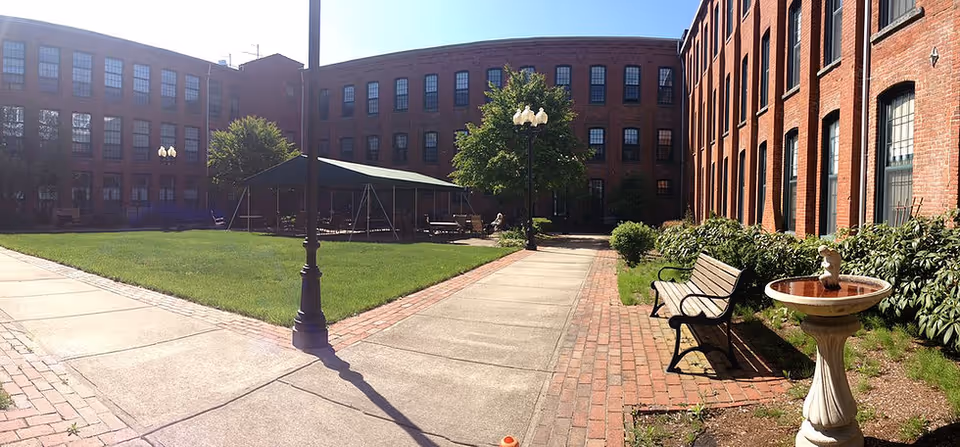 Outdoor courtyard area of Franklin Court Assisted Living with brick buildings surrounding a grassy lawn. There are paved walkways, benches, a birdbath, and a covered seating area with tables and chairs. Trees and shrubs are planted along the walkways.