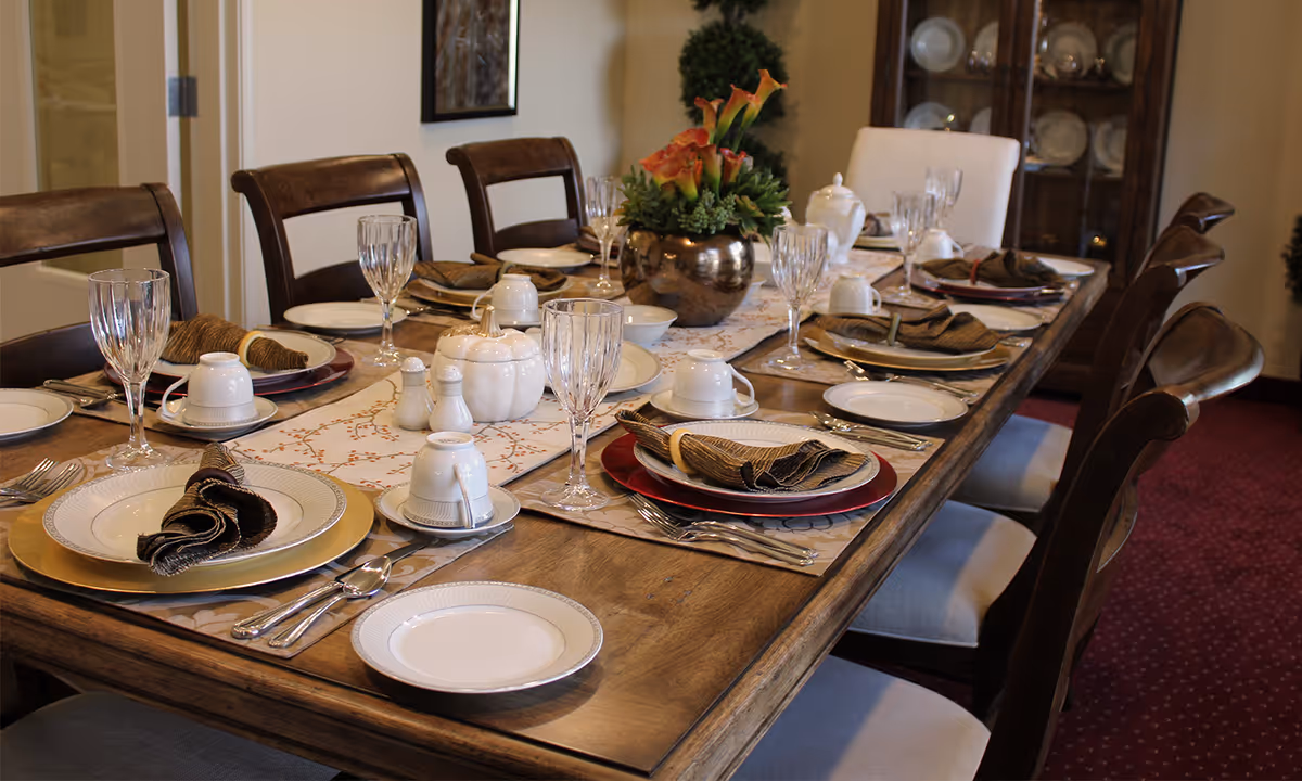 A dining table set for a meal with eight place settings including plates, glasses, cups, silverware, and folded napkins. The table has a decorative runner and a centerpiece with flowers. The room has wooden chairs and a cabinet with dishes in the background.