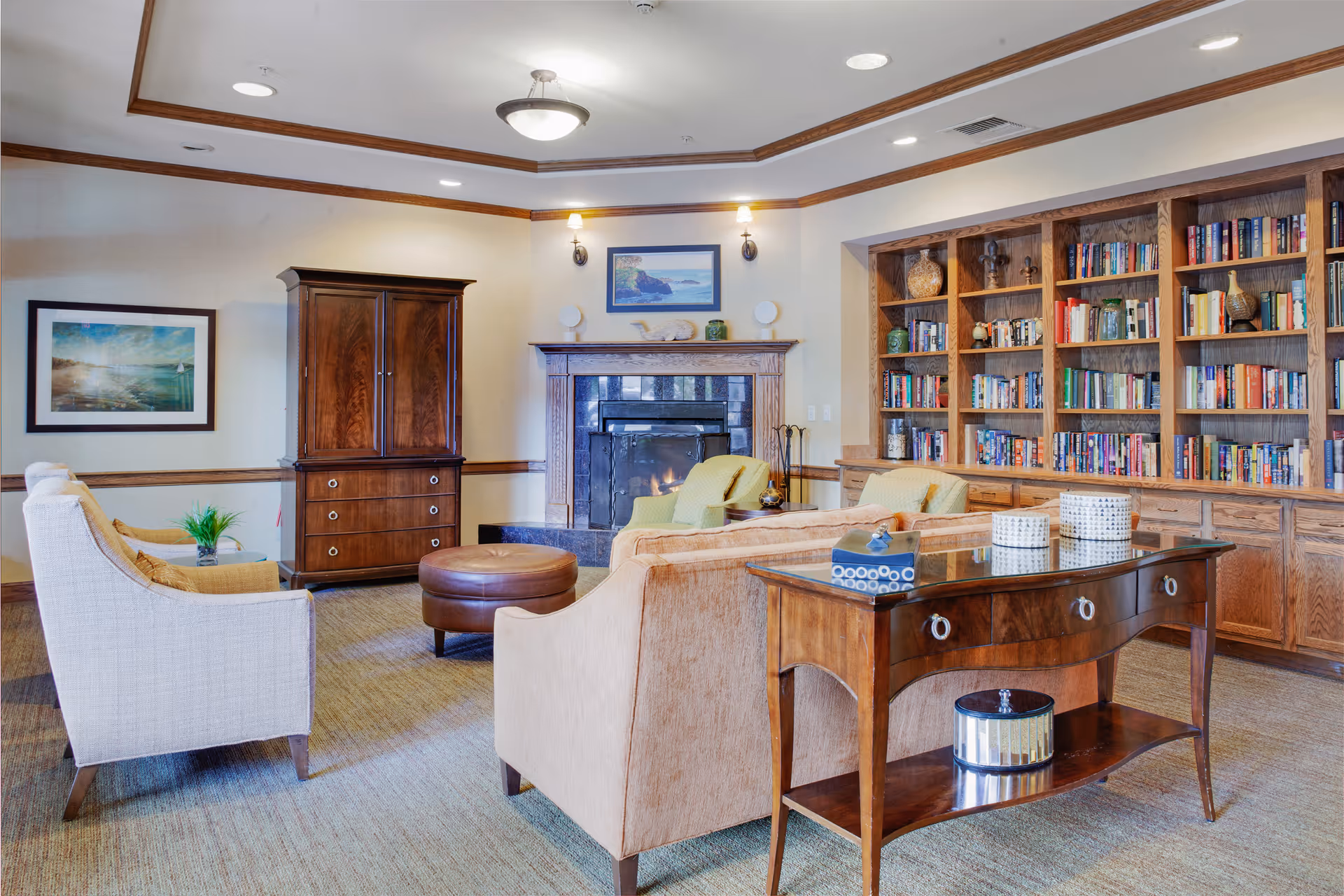 A cozy living room area in a senior living facility featuring a fireplace with a mantle, two armchairs, a beige sofa, a round leather ottoman, a wooden cabinet, and a large built-in bookshelf filled with books and decorative items. The room has soft lighting with wall sconces and a ceiling light, beige walls with wooden trim, and carpeted flooring.